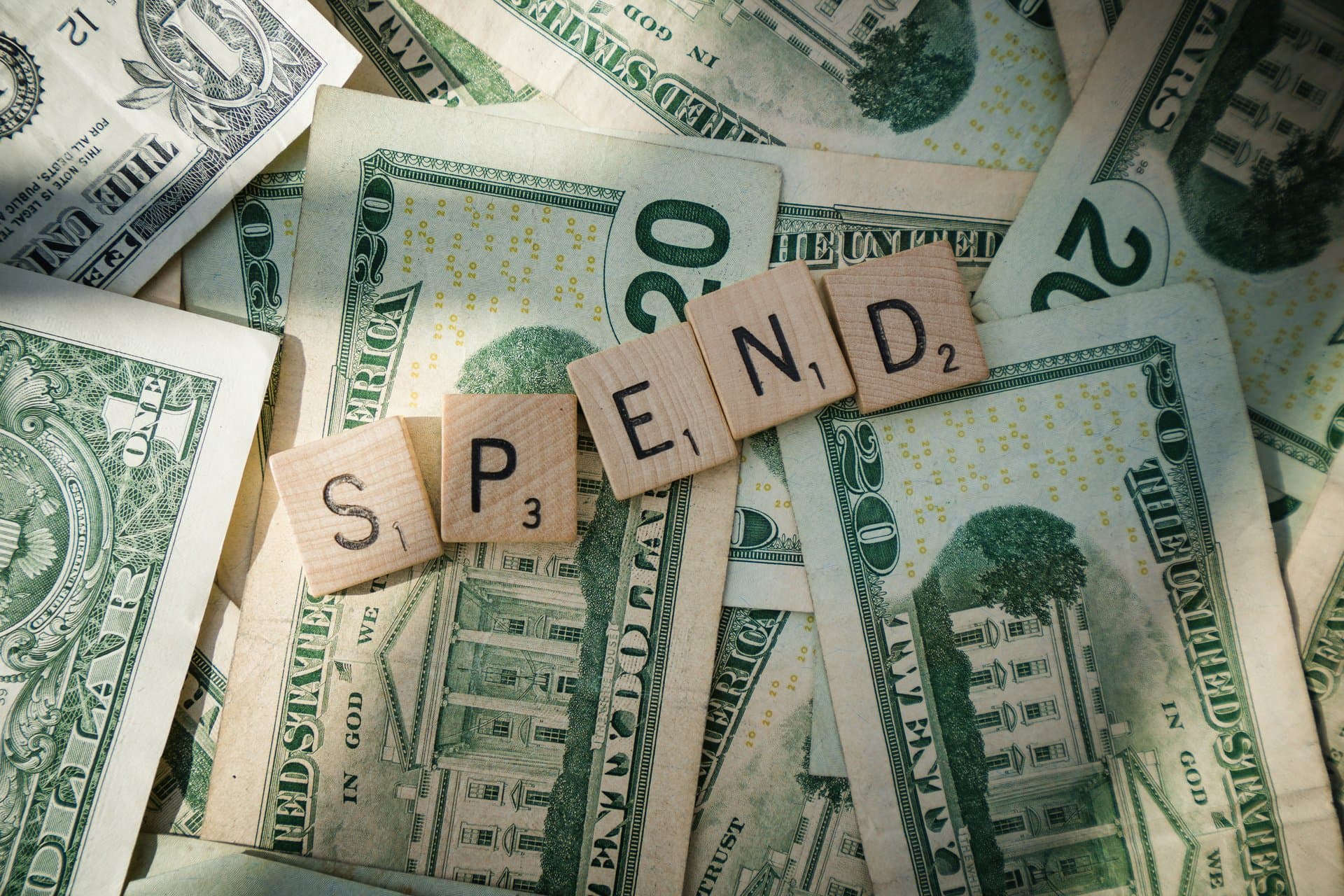 a scrabble tile with spend on top of a spread of dollars